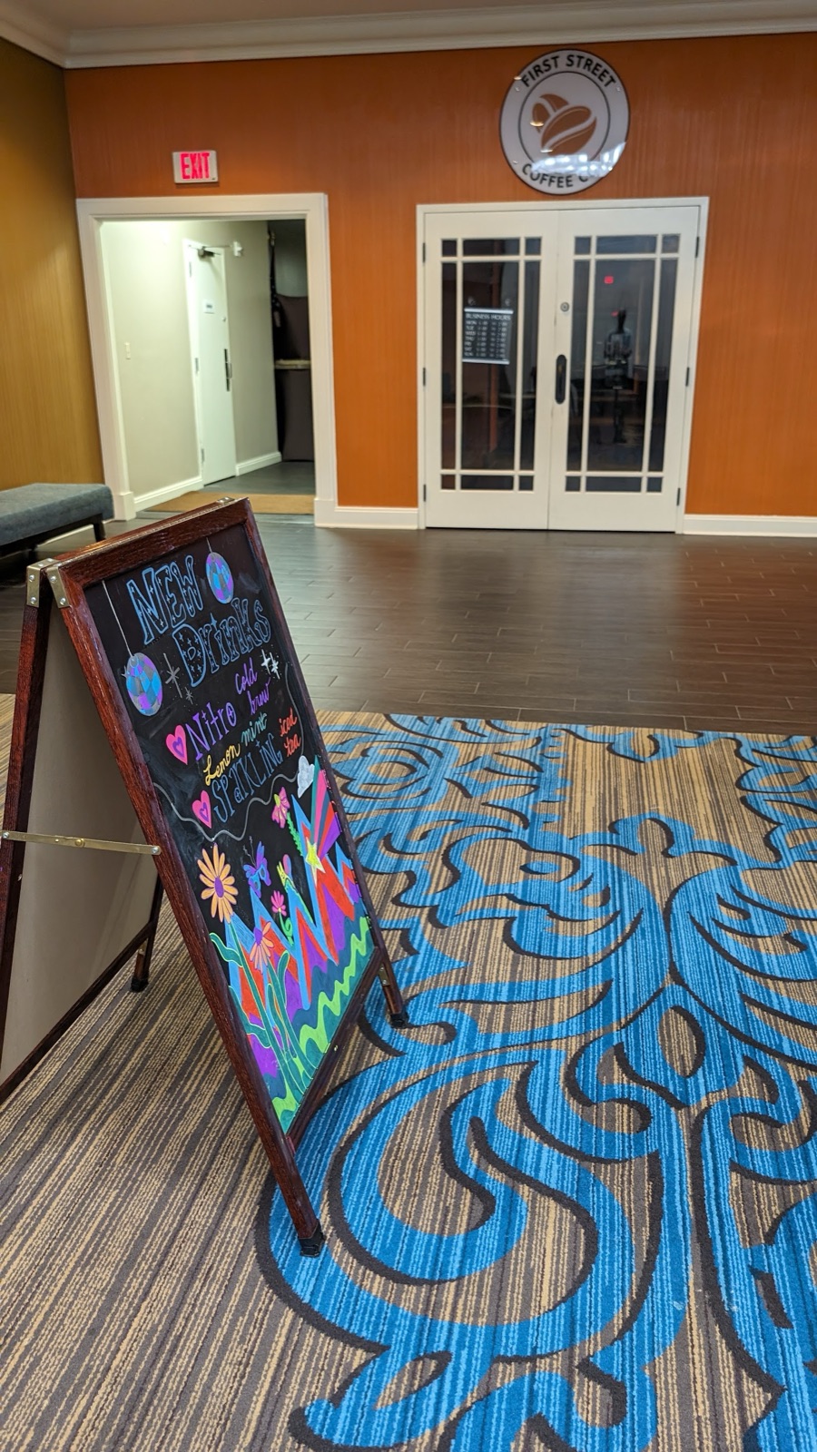 The entrance to First Street Coffee: an orange wall with a round sign, off the hotel's blue patterned carpet.
