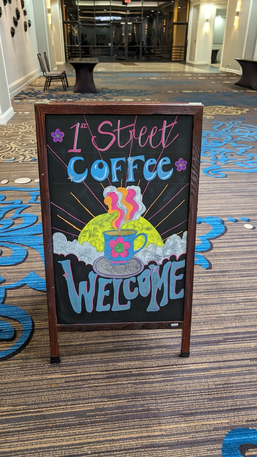 A hand-lettered chalkboard sign reading First Street Coffee Welcome, with a drawing of a steaming coffee cup.